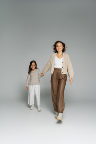 happy and stylish woman looking at camera while holding hands with daughter and walking on grey background