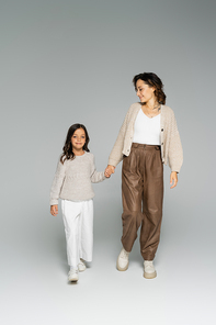smiling woman in stylish autumn clothes walking with daughter on grey background