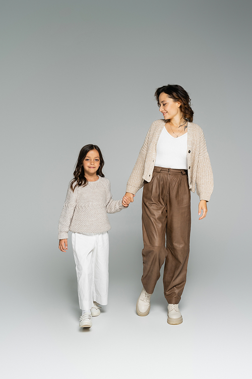 smiling woman in stylish autumn clothes walking with daughter on grey background