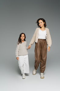 mother with child in stylish autumn outfit holding hands and looking at camera while walking on grey background