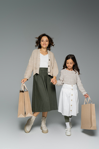full length of mom and daughter in skirts holding hands while carrying shopping bags on grey background