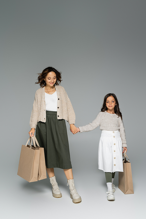 full length of woman and child in knitwear and skirts holding hands and walking with shopping bags on grey background