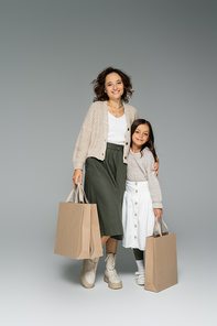 happy woman in stylish autumn clothes holding shopping bags and hugging daughter on grey background