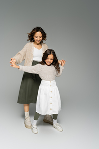 happy mother and child in stylish clothes holding hands and dancing on grey background
