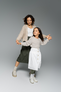 happy girl and woman in trendy clothes dancing while holding hands on grey background