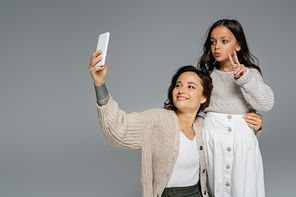 trendy girl showing victory sign near smiling mom taking selfie on cellphone isolated on grey