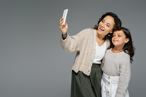 excited woman taking selfie on smartphone with daughter in trendy clothes isolated on grey
