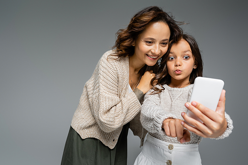 amazed girl pointing at mobile phone near smiling mother in knitwear isolated on grey