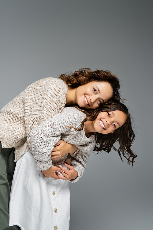 cheerful woman and child in warm outfit embracing and smiling at camera isolated on grey
