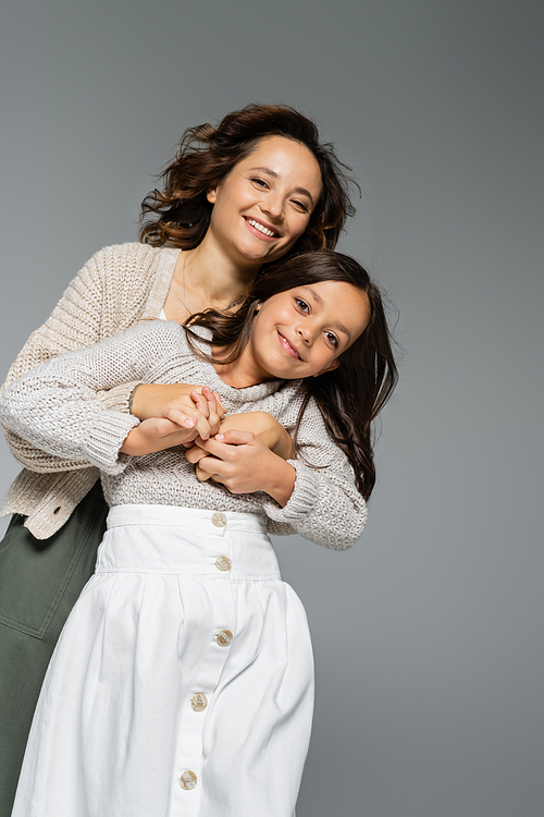 low angle view of happy young woman in knitwear hugging smiling daughter in skirt isolated on grey