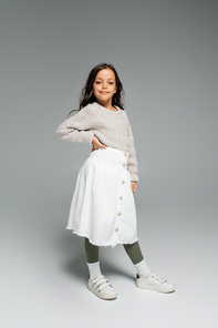 full length of child in white skirt and knitted sweater standing with hand on hip on grey