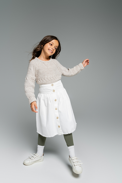 full length of happy girl in warm sweater and white skirt posing on grey background