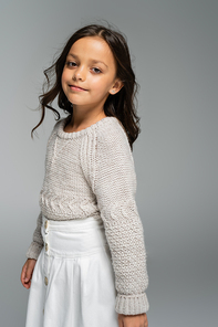 smiling girl in warm sweater and white skirt looking at camera isolated on grey