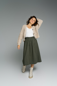 full length of trendy woman in knitted cardigan and skirt with boots standing with hand behind head on grey