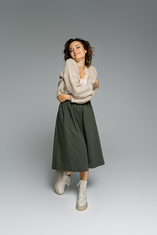 full length of pleased woman in warm cardigan and green skirt posing on grey