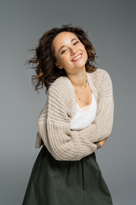 joyful woman in knitted cardigan and green skirt posing with crossed arms isolated on grey