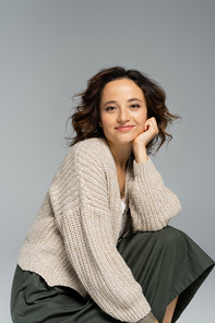 brunette woman in cozy cardigan and green skirt holding hand near face while smiling at camera isolated on grey