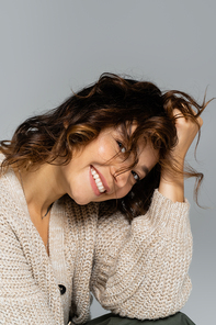 happy woman in knitted cardigan touching wavy hair and looking at camera isolated on grey