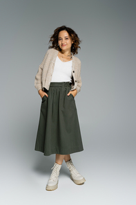 full length of young woman in cozy cardigan and boots posing with hands in pockets of green skirt on grey background
