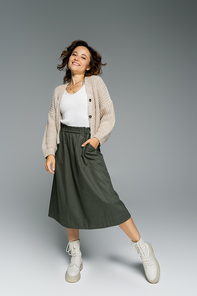 cheerful woman in warm cardigan and boots posing with hand in pocket of green skirt on grey background