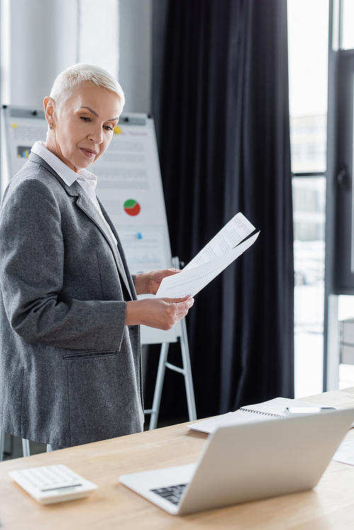 stylish businesswoman standing with documents at workplace during online conference on laptop