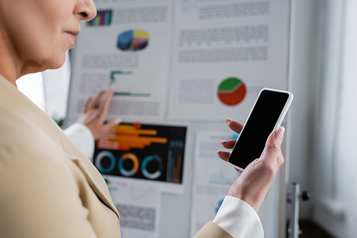 cropped view of banker holding smartphone with blank screen near blurred flip chart with graphs