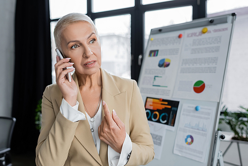 stylish businesswoman talking on mobile phone near flip chart on blurred background