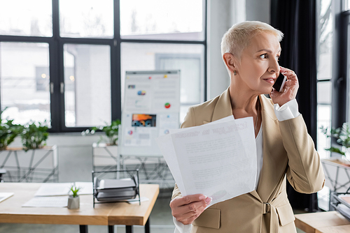 stylish banker with documents looking away while talking on smartphone in blurred office