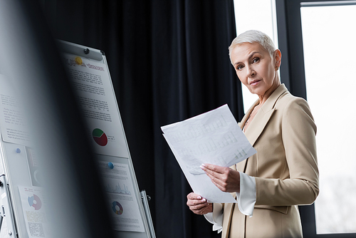 senior businesswoman with papers looking at camera near flip chart with infographics