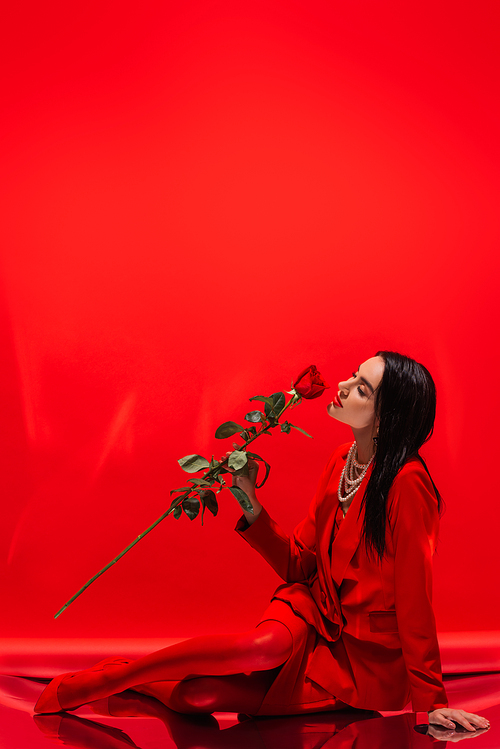 Pretty brunette woman in jacket smelling rose while sitting on red background