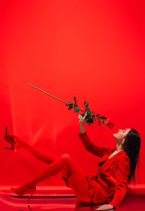 Side view of brunette model in heels smelling rose while sitting on red background
