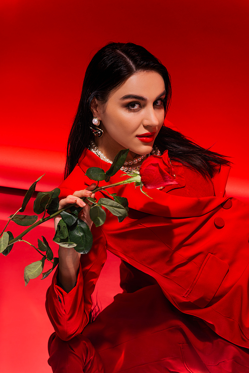 Pretty brunette woman in jacket holding rose and looking at camera on red background