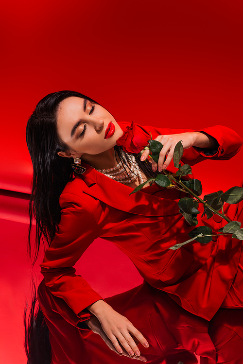 Young woman in jacket and pearl necklace holding rose and closing eyes on red background