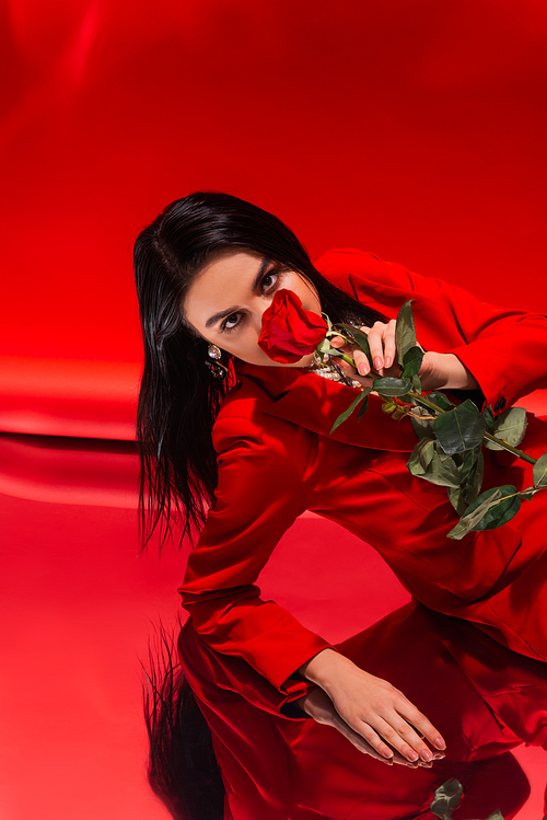 Brunette woman in jacket covering face with rose on reflective surface on red background