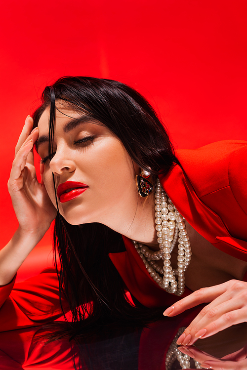 Portrait of elegant brunette woman in pearl necklace lying on reflective surface isolated on red