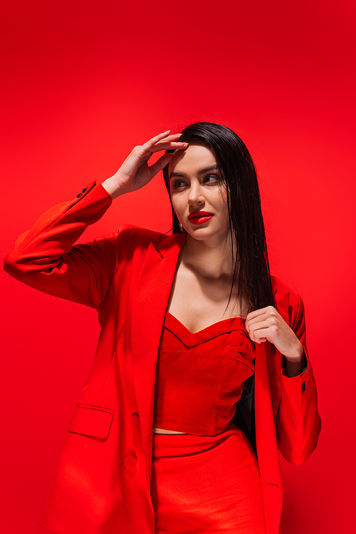 Stylish brunette woman in jacket posing and looking away isolated on red