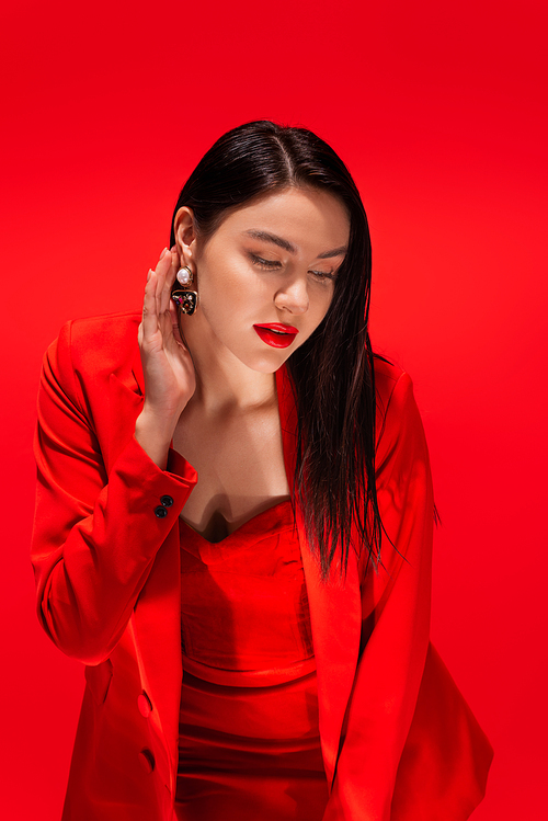 Elegant model in jacket touching earring while posing isolated on red