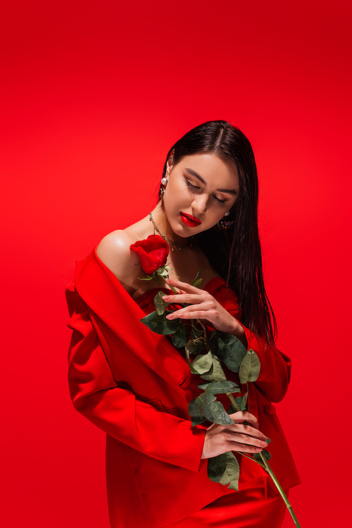 Stylish brunette model in jacket holding rose while closing eyes isolated on red