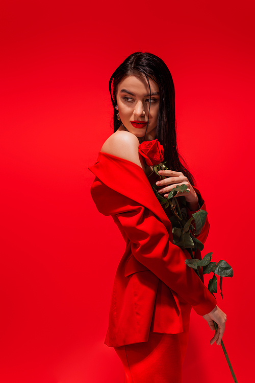 Elegant brunette woman in jacket with naked shoulder holding rose isolated on red