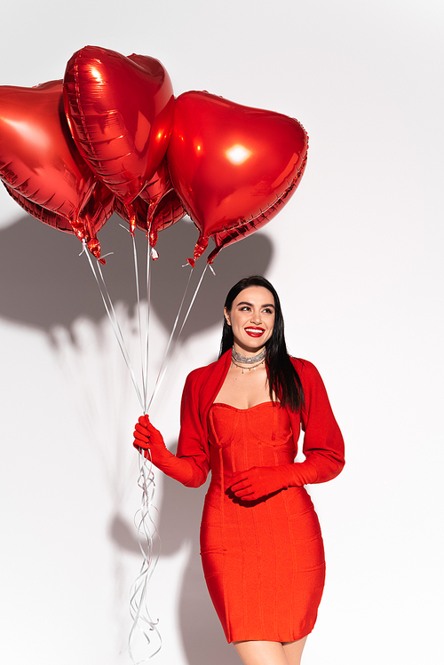 Cheerful brunette woman holding red heart shaped balloons on white background