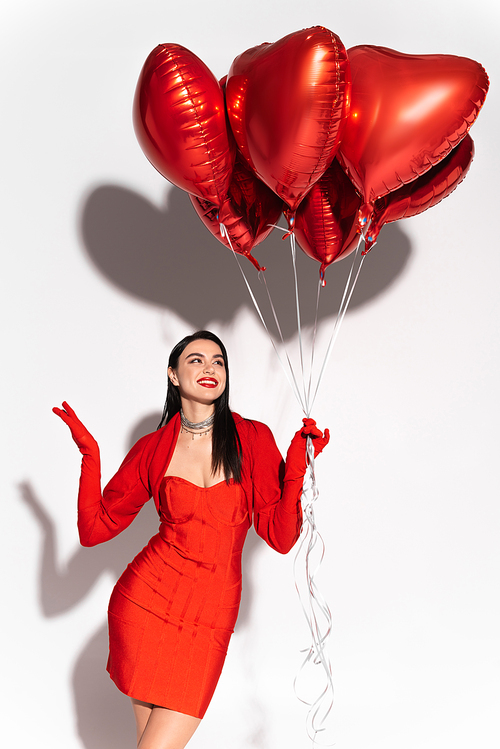 Positive and stylish woman in red clothes looking at heart shaped balloons on white background with shadow