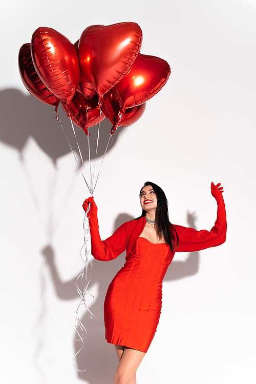 Cheerful brunette woman in red dress looking at heart shaped balloons on white background with shadow