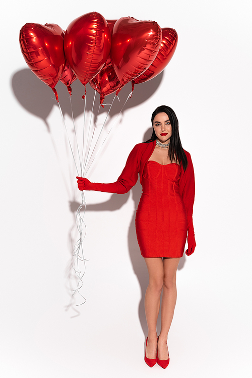 Full length of stylish woman in red dress and heels holding heart shaped balloons on white background with shadow