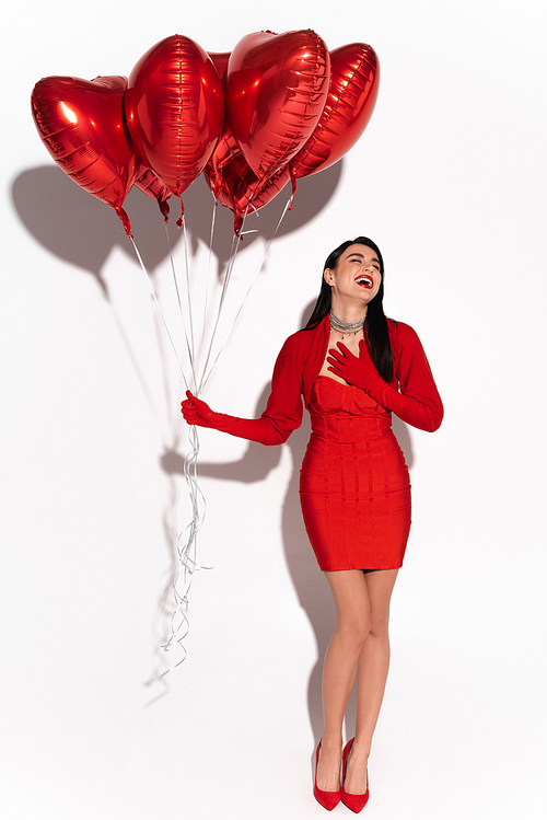 Stylish woman in red clothes laughing and holding heart shaped balloons on white background with shadow