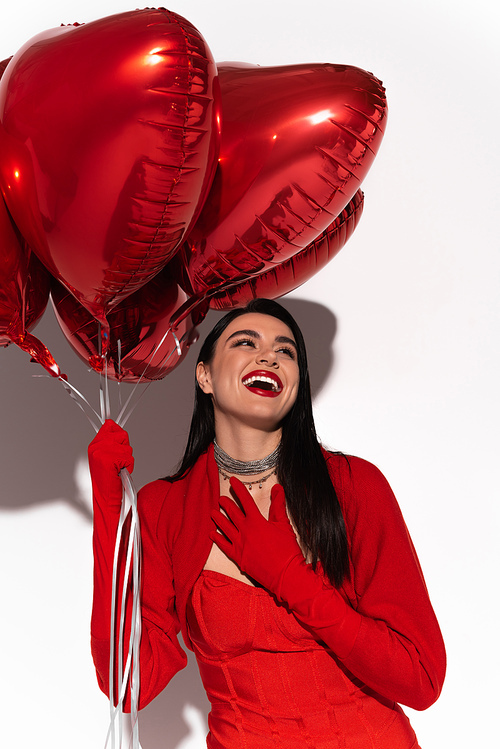 Excited brunette woman in red jacket holding heart shaped balloons on white background with shadow