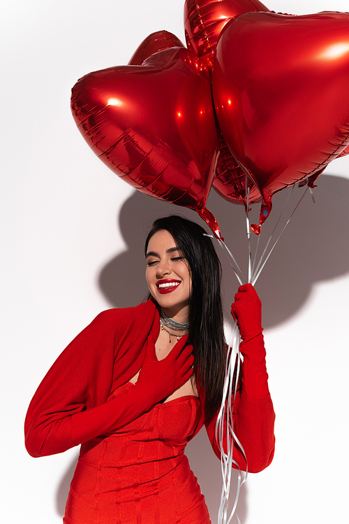 Positive brunette woman in red dress posing with heart shaped balloons on white background with shadow
