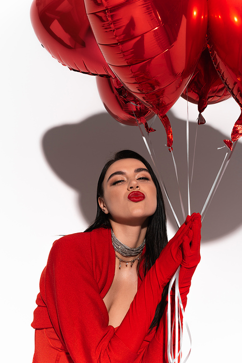Stylish brunette woman in red gloves holding heart shaped balloons and blowing air kiss on white background with shadow