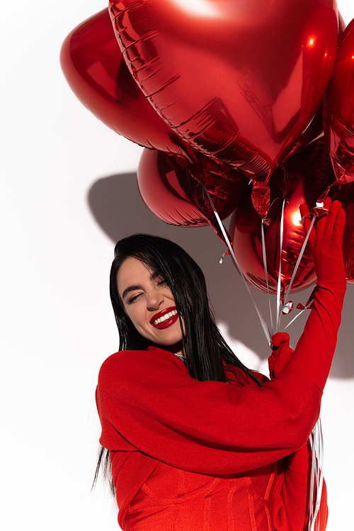 Happy brunette woman in red jacket touching heart shaped balloons on white background with shadow