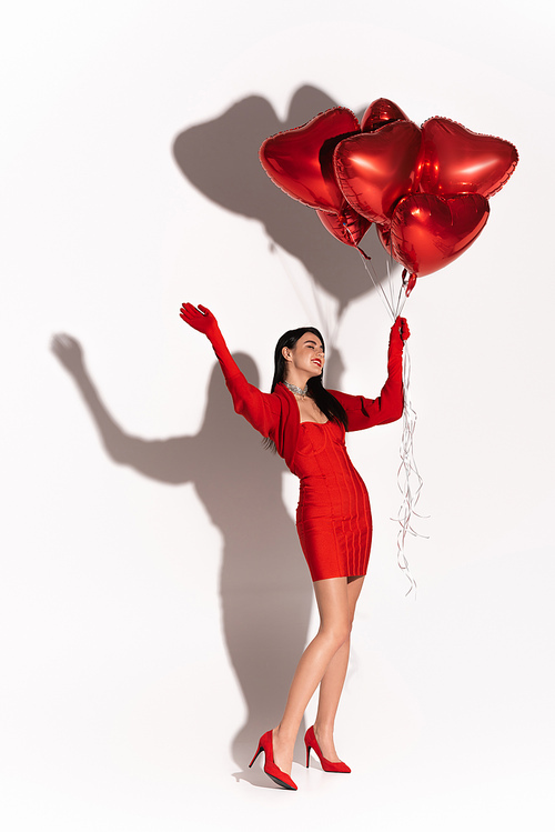 Cheerful woman in heels and dress holding red heart shaped balloons and waving hand on white background with shadow