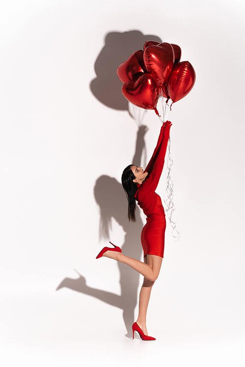 Excited young woman in red clothes and heels holding heart shaped balloons on white background with shadow